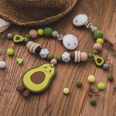 Avocado-shaped baby teething toy with silicone beads and pacifier clip on wooden surface.