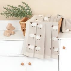 Soft beige baby blanket with cute white and brown sheep pattern, draped over a white dresser with wooden knobs, next to a small wooden teddy bear and eucalyptus plant in a wicker basket, ideal for nursery decor.