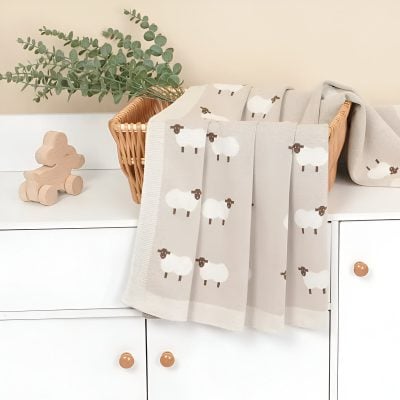 Soft beige baby blanket with cute white and brown sheep pattern, draped over a white dresser with wooden knobs, next to a small wooden teddy bear and eucalyptus plant in a wicker basket, ideal for nursery decor.