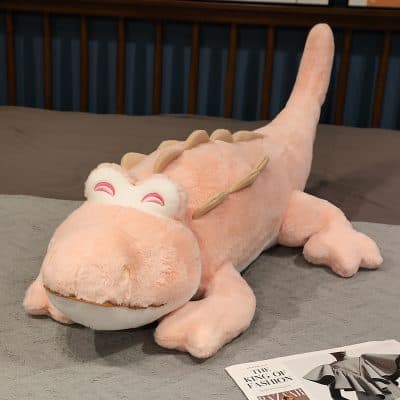 Soft pink plush dragon stuffed toy with smiling eyes, lying on a bed; perfect as a cozy decor or playful gift for children and dragon lovers.