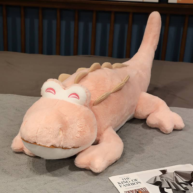 Soft pink plush dragon stuffed toy with smiling eyes, lying on a bed; perfect as a cozy decor or playful gift for children and dragon lovers.