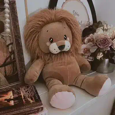 Soft plush lion toy sitting on a white shelf, surrounded by decorative items including books, flowers, and framed prints, in a cozy home decor setting.