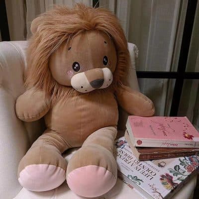 Soft plush lion stuffed animal sitting on a white armchair near a stack of colorful books, adding a cozy and playful touch to the room decor for children's comfort and home aesthetics.