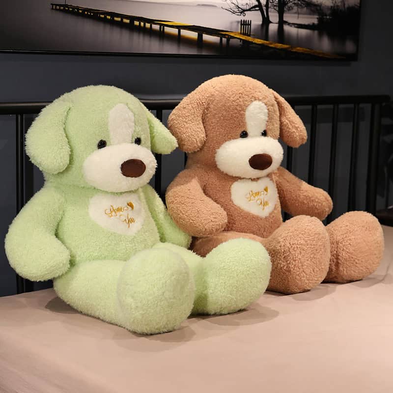 Soft plush teddy bears in light green and brown colors, perfect for gifts, with embroidered "Love You" messages on their chests, displayed on a bed in a cozy indoor setting.
