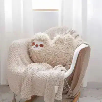 Soft beige plush sloth pillow resting on a cozy textured blanket, placed on an ergonomic white chair near sheer white curtains, emphasizing comfort and relaxation.