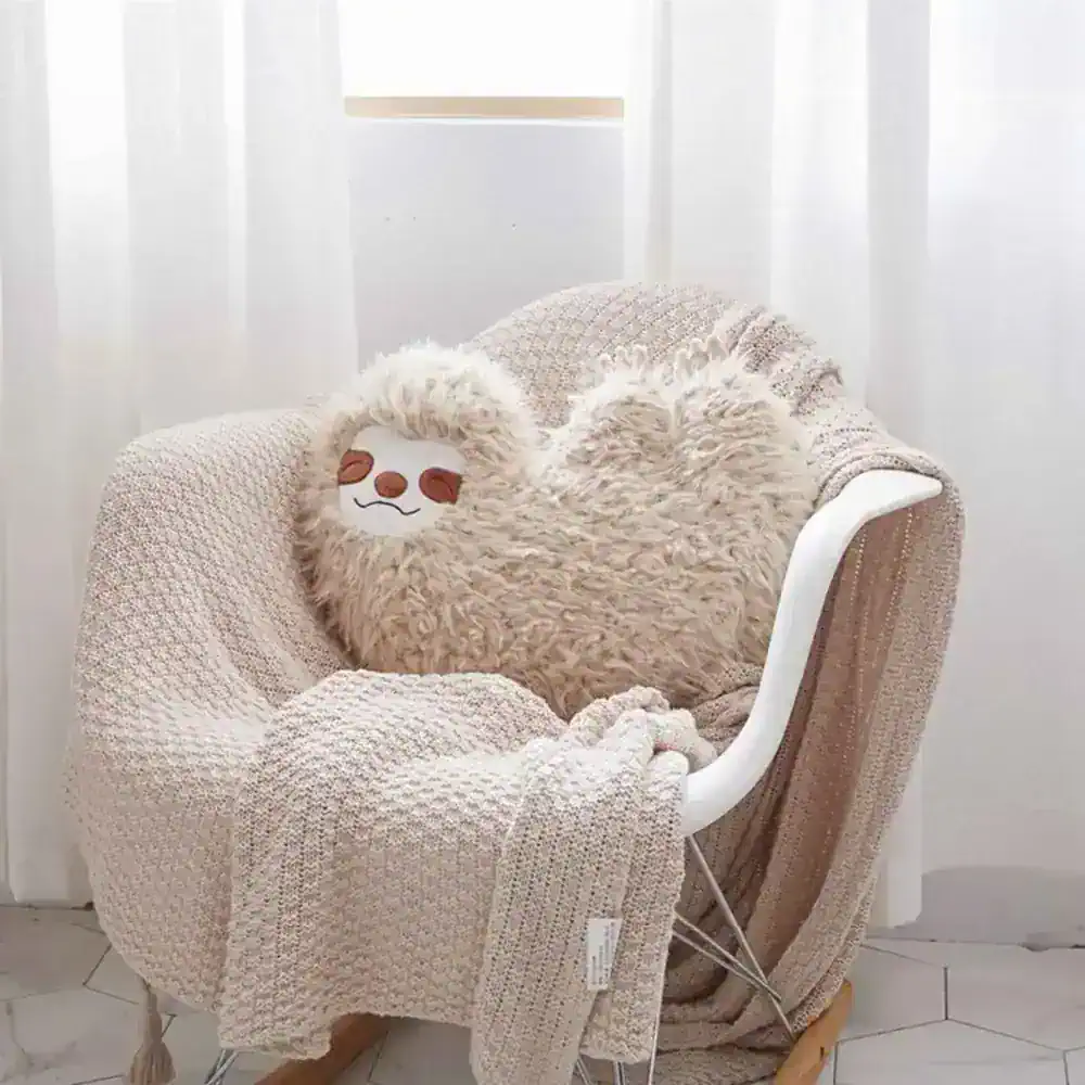 Soft beige plush sloth pillow resting on a cozy textured blanket, placed on an ergonomic white chair near sheer white curtains, emphasizing comfort and relaxation.