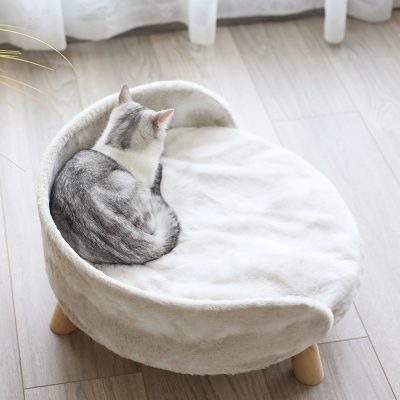 Cozy gray and white cat curled up on a plush, cream-colored pet bed with wooden legs, in a bright, modern living space. Perfect for pet comfort and relaxation.