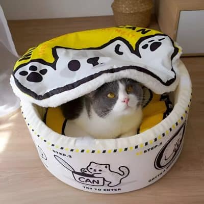 Fluffy gray and white cat lying inside a round, cozy cat bed with a cartoon cat design and yellow interior, partially covered by a plush blanket, on a wooden floor.