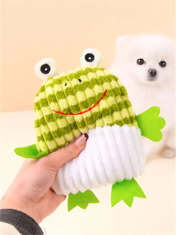 Bright green and white plush frog toy with googly eyes, smiling face, and felt limbs, held in a person's hand with adorable white puppy in the background.