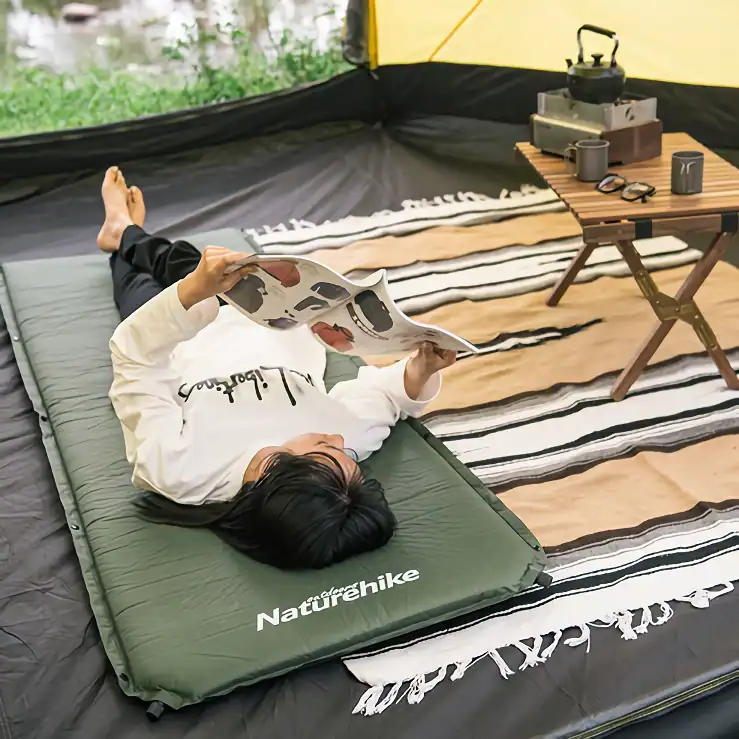 Relaxing camping setup with a person reading a magazine inside a tent, outdoor adventure gear, portable camping mattress, and a wooden table with kettle and mugs for outdoor leisure and exploration.