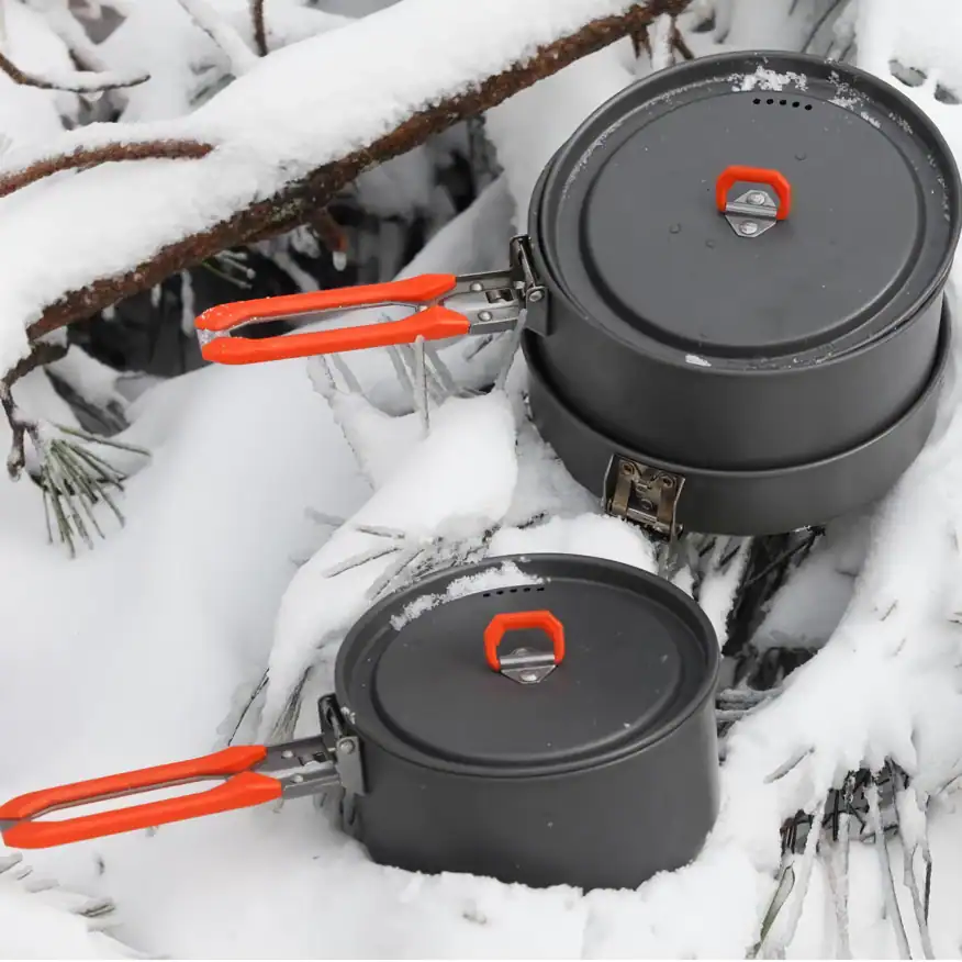 Cookware set with insulated, heat-resistant lids and bright orange handles, designed for outdoor winter camping in snow. Durable construction ideal for outdoor cooking.