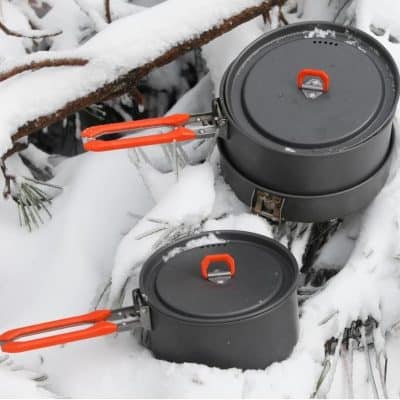 Camping cookware set in snow with black pots and orange handles, outdoor winter cooking tools.