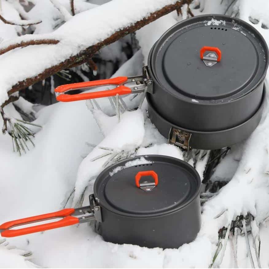 Camping cookware set in snow with black pots and orange handles, outdoor winter cooking tools.