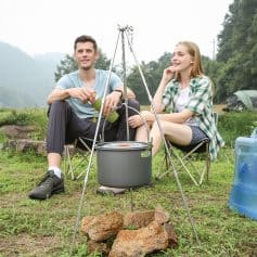 Outdoor camping scene with two young adults sitting by a portable stove, enjoying a meal in nature, surrounded by greenery and camping gear, promoting outdoor adventure and travel.