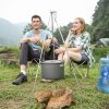 Outdoor camping cooking setup with portable pot over fire ring, friends enjoying meal outdoors, nature camping gear, mountains, and forest background.
