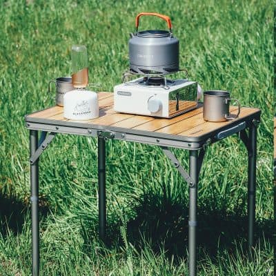 Compact portable camping stove with cookware and mugs set on a wooden table outdoors.
