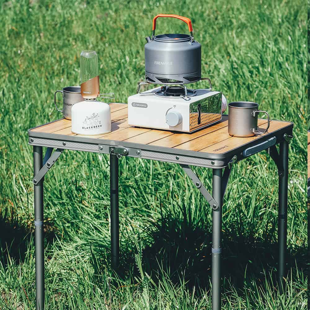 Compact portable camping stove with cookware and mugs set on a wooden table outdoors.
