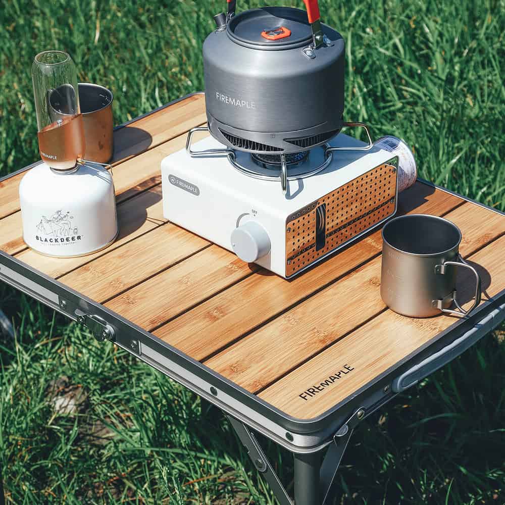Portable camping stove setup for outdoor cooking with Firemaple and Blackdeer gear, including a kettle and mugs, on a wooden table in nature.