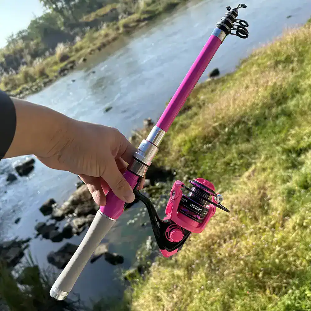 Bright pink fishing rod and reel near riverbank, outdoor fishing scenery with flowing water and rocks, ideal for fishing enthusiasts seeking quality fishing gear.