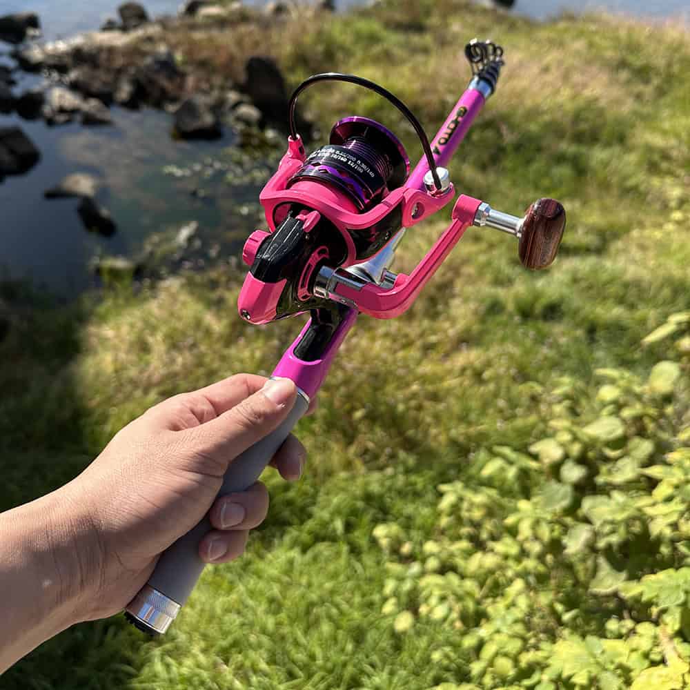 Pink fishing reel and rod set outdoors by a river, emphasizing fishing gear quality.