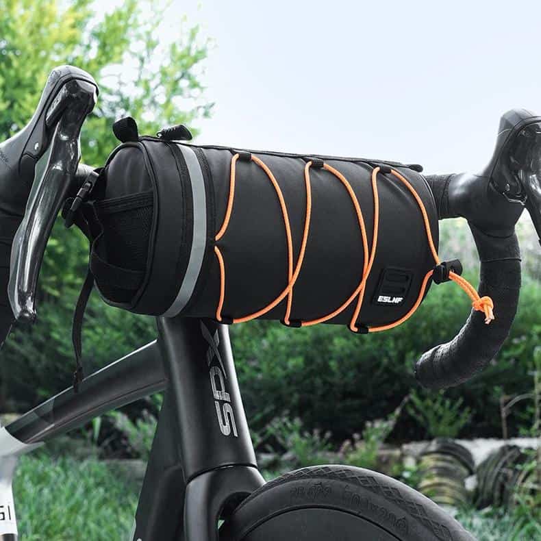 Waterproof bicycle handlebar bag for outdoor cycling accessories, black with orange bungee cords, designed for bike storage and organization, shown mounted on a bike handlebar outside in nature.