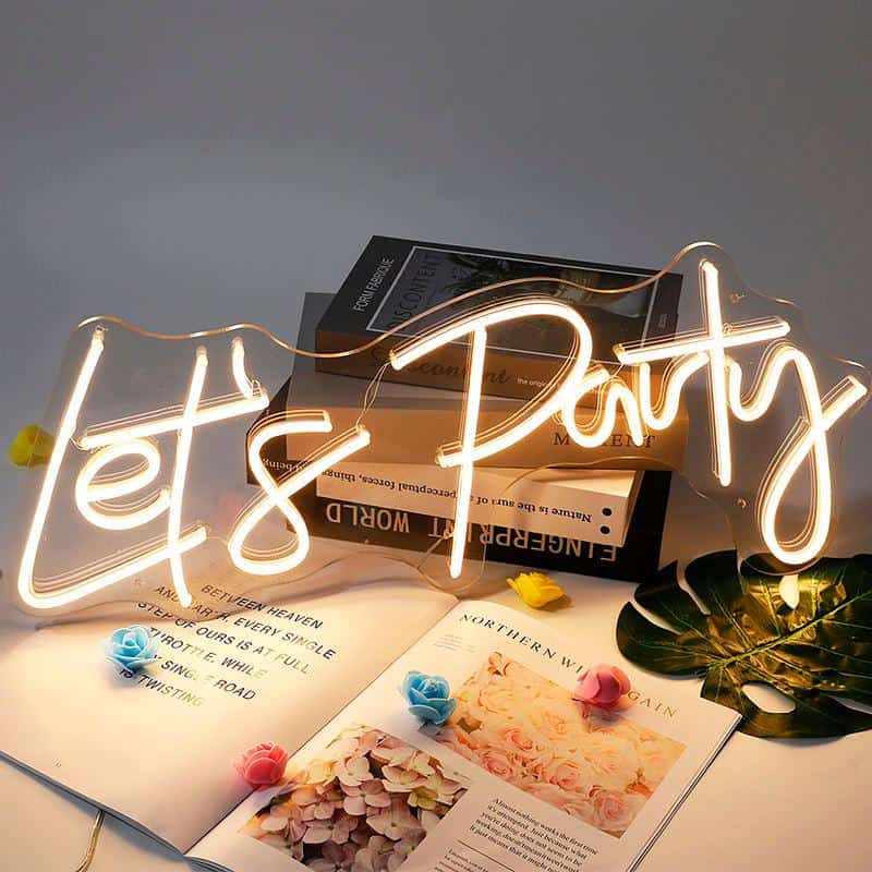 Neon "Let's Party" sign illuminated over vanity table with fashion and lifestyle magazines, floral accents, and tropical leaves, vibrant party decor for celebrations, stylish ambiance for special occasions.