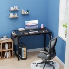 Modern home office setup with black desk, ergonomic gray chair, desktop computer, bookshelf, and decorative wall shelves, creating a stylish and functional workspace.