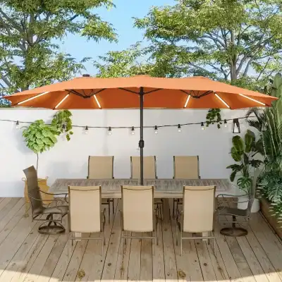 Outdoor dining area with a large tan umbrella, wooden table, and multiple beige chairs, surrounded by lush green plants and string lights, perfect for a relaxing meal or gathering.