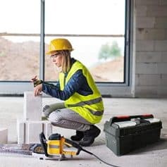 Detailed description of a female construction worker wearing a safety helmet and high-visibility vest working on bricks with tools at a building site.