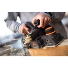 Alt text: Close-up of hands operating a cordless sander peeling wood, demonstrating its use in woodworking, DIY, and home improvement projects.