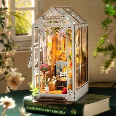 A detailed miniature greenhouse model decorated with vibrant flowers and cozy furniture, placed on a large book in a bright, welcoming space. Perfect for hobbyists and home decor enthusiasts.