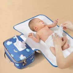 Baby lying on changing pad being dressed with diaper, medical supplies and portable diaper bag nearby.