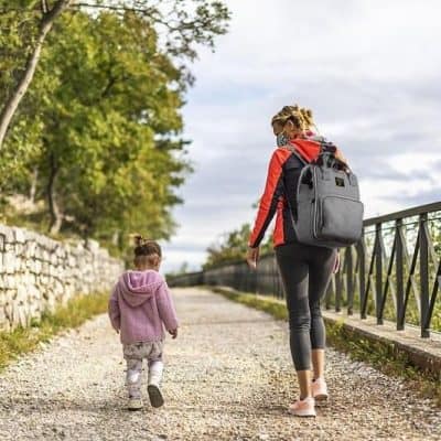 A mother and young girl walking outdoors along a scenic pathway with backpacks, enjoying a peaceful day. Perfect for those looking for stylish, comfortable backpacks from Monalisa Store for family adventures.