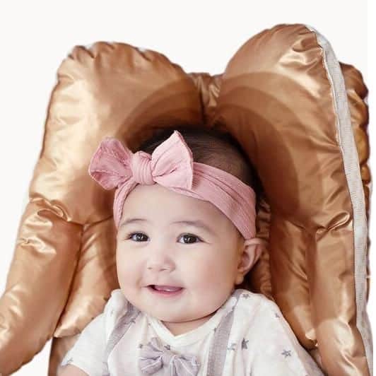 Adorable baby girl with a pink headband and bow, seated in a plush, satin-like baby chair. Perfect for baby care accessories, baby chairs, and infant comfort products.