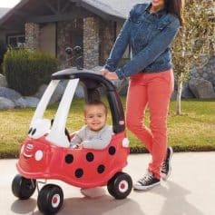 Bright-eyed toddler in a red ladybug stroller with an adult outdoors, sunny day, lush yard background. Child wears a gray outfit, joyful expressions. Emphasizes playful, safe kids’ eyewear and optical products at MonaLisa Store.