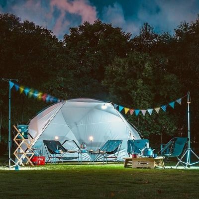 A beautifully lit outdoor camping scene featuring a white tent, colorful string lights, camping chairs, and accessories set in a forest clearing during evening hours.