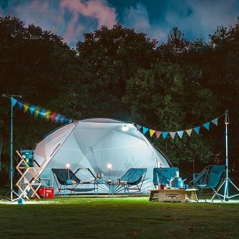 A beautifully lit outdoor camping scene featuring a white tent, colorful string lights, camping chairs, and accessories set in a forest clearing during evening hours.