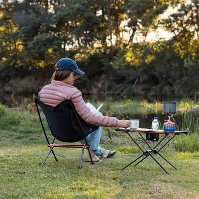 Relaxing woman reading outdoors by a lake, enjoying nature with camping gear, perfect for outdoor leisure and camping accessories.