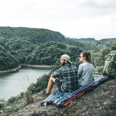 A couple sitting on a portable sleeping pad overlooking a serene river surrounded by lush green hills, enjoying outdoor exploration and nature. Perfect for outdoor lovers and hikers.
