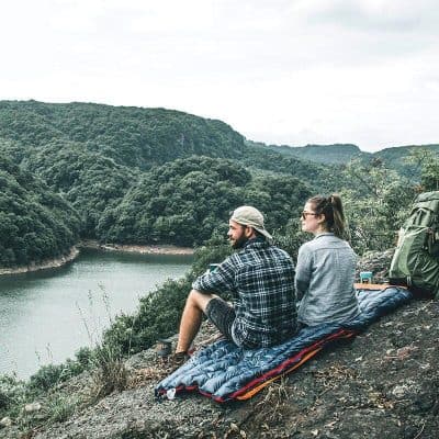 A couple sitting on a portable sleeping pad overlooking a serene river surrounded by lush green hills, enjoying outdoor exploration and nature. Perfect for outdoor lovers and hikers.
