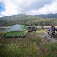 Great outdoor camping scene near a serene lake, featuring a green tent, camping chairs, and a woman relaxing by a picnic table with beautiful mountain views. Perfect for nature exploration, hiking, and outdoor adventures with friends or family.