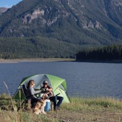 A couple enjoying camping near a serene lake with a green tent and a dog, set against beautiful mountain scenery, ideal for outdoor enthusiasts seeking adventure and relaxation.