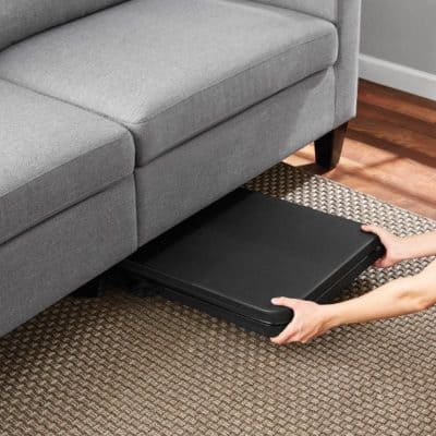 A person is placing a black footrest or ottoman in front of a cozy, light gray sofa in a modern living room. The setting features warm wooden flooring and an accent rug, perfect for relaxing and enhancing living space aesthetics.