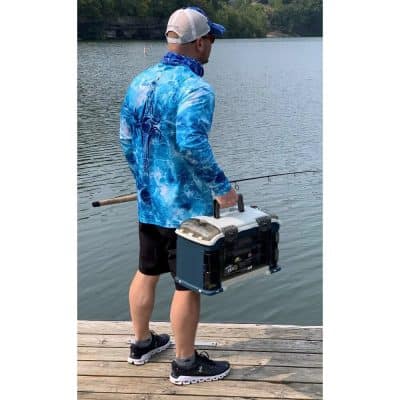 A man dressed in a vibrant bluewater-themed jacket fishing by a serene lake, carrying a compact tackle box, perfect for outdoor fishing adventures.