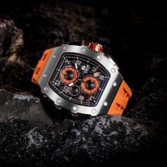 A high-end luxury sports watch with a bold orange strap, intricate chronograph dials, and a robust stainless steel case, placed on dark volcanic rocks. Designed for stylish men seeking durable, fashionable watches for active and outdoor use.