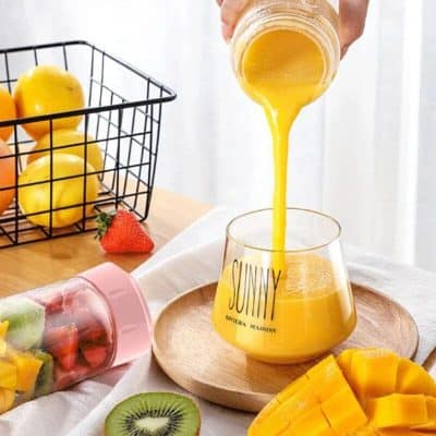 Bright yellow fruit juice being poured into a glass, surrounded by fresh fruits like strawberries, kiwi, and mango, with a basket of lemons in the background.