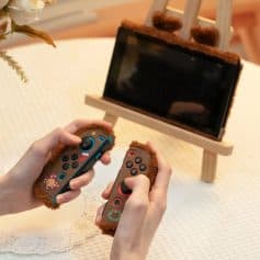 Close-up of hands holding cute, furry Nintendo Switch controllers decorated with holiday-themed designs, showing a playful and cozy gaming setup on a white lace tablecloth with a wooden easel in the background.