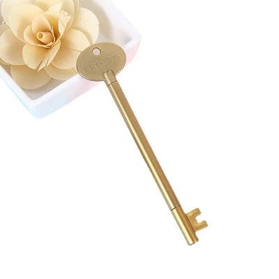 A gold-colored antique-style key resting beside a white flower in a small container. Perfect for vintage door decor or as a decorative key. Elegant and timeless design for home or office.
