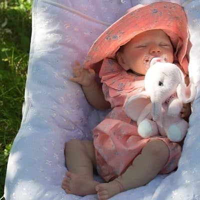 Adorable baby with elephant stuffed animal, wearing cute pink outfit and sunhat, resting on a cozy white cushion outdoors. Perfect for baby care and gift ideas.