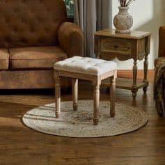 Vintage-style wooden vanity stool with tufted white cushion, placed on a decorative rug in a cozy, well-decorated living room. Perfect for bedroom or dressing room decor.